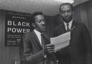 Two CORE officials discuss a memo while standing in front of a sign in their office that reads "Support Black Power"
