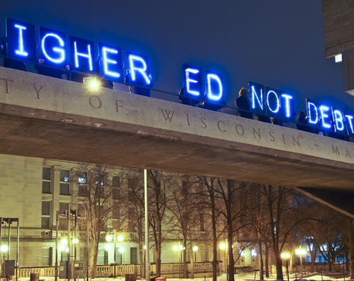 A glowing sign reading "Higher Ed Not Debt" above a university campus