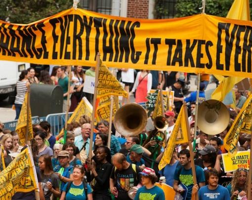 Marchers beneath a banner that reads "To Change Everything It Takes Everyone"