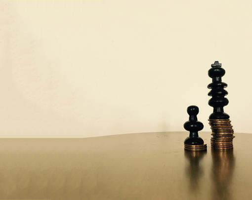 Two chess pieces, a king and a pawn, atop unequal stacks of coins.
