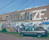 a wall is half covered in graffiti, in the top half, which is mostly unadorned, the word 'DECOLONIZE' is painted in large black and white letters