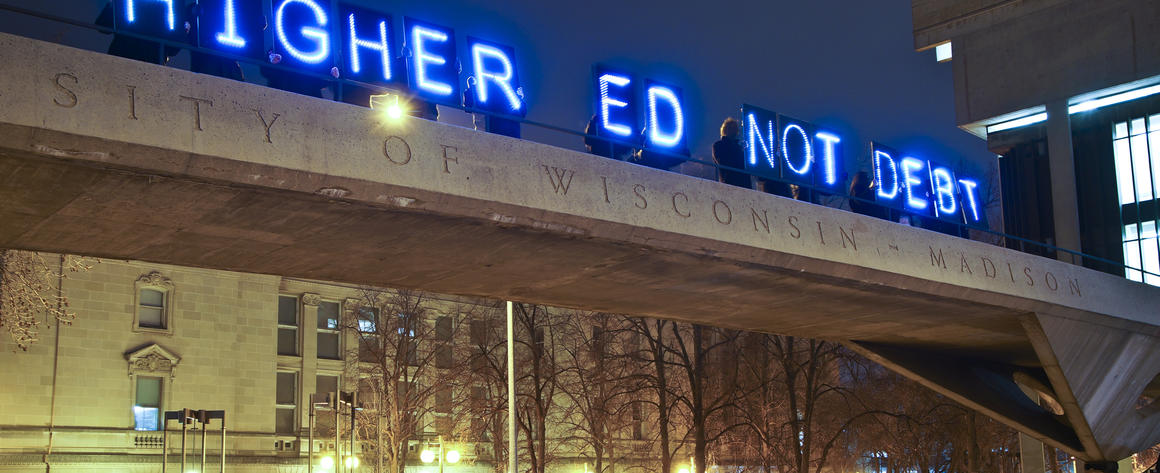 A glowing sign reading "Higher Ed Not Debt" above a university campus