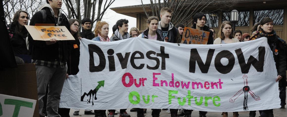 Students standing behind a banner that reads "Divest Now"