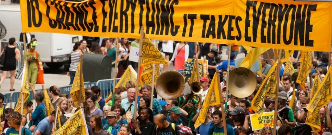 Marchers beneath a banner that reads "To Change Everything It Takes Everyone"